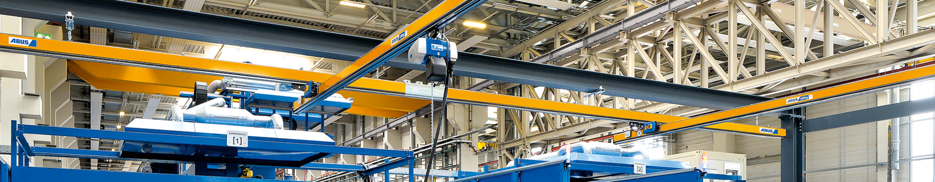 Jednobryłowa kolejka podwieszana typu EHB W hali przemysłowej widocznych jest kilka jednobryłowych kolejek podwieszanych typu EHB. Na pierwszym planie pracownik obsługujący wciągnik łańcuchowy za pomocą wiszącego przycisku. Wciągnik łańcuchowy porusza się za pomocą wózka rolkowego w profilu głównej belki nośnej.