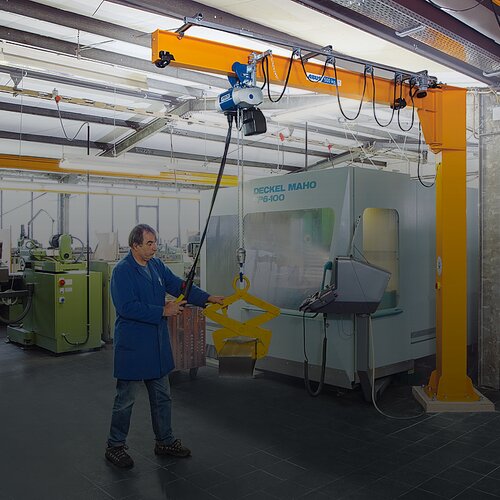 Pracownik obsługuje suwnicę kolumnową i podnosi ładunek za pomocą wciągnika łańcuchowego w hali produkcyjnej.