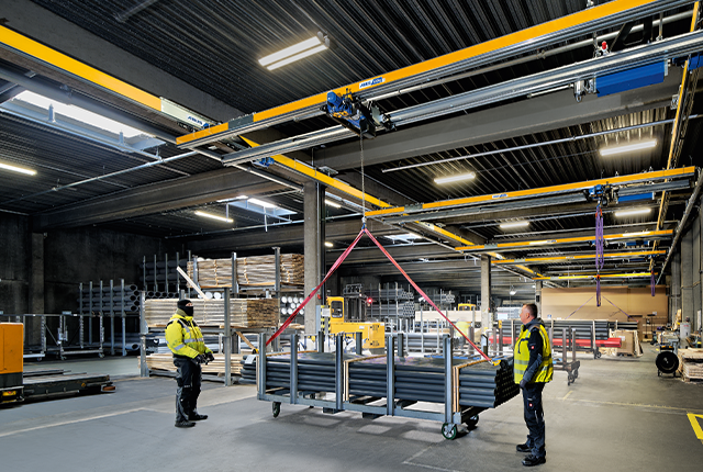 Suwnica dwudźwigarowa ZHB przeprowadza podnoszenie ładunku w hali przemysłowej, podczas gdy pracownicy nadzorują tę czynność.