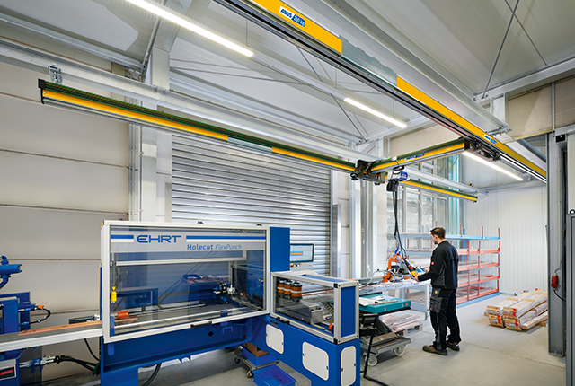 Suwnica jednodźwigarowa EHB-X przemieszcza ładunek w hali przemysłowej, podczas gdy w tle pracownik pracuje przy maszynie.