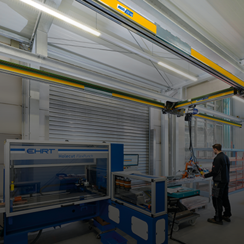 Suwnica jednodźwigarowa EHB-X przemieszcza ładunek w hali przemysłowej, podczas gdy w tle pracownik pracuje przy maszynie.